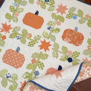 Pode incluir: Uma colcha branca com um design de patchwork que apresenta abóboras laranja, folhas verdes e estrelas azuis e brancas. A colcha é feita com uma variedade de tecidos e tem uma textura acolchoada.