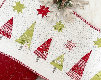 May include: A white quilted table runner with a red and green Christmas tree design. The runner features a repeating pattern of trees, stars, and snowflakes. The runner is trimmed with a red and white polka dot border.