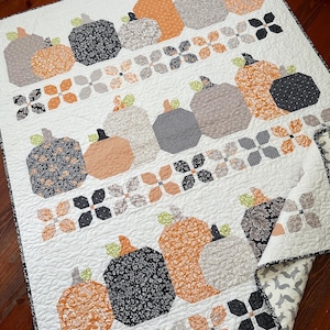 May include: A white quilt with a patchwork design featuring pumpkins in various shades of orange, gray, and black. The pumpkins are arranged in rows and are surrounded by a pattern of small, white flowers.