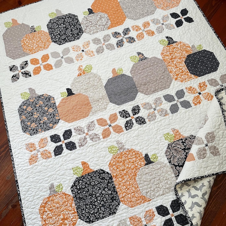May include: A white quilt with a patchwork design featuring pumpkins in various shades of orange, gray, and black. The pumpkins are arranged in rows and are surrounded by a pattern of small, white flowers.