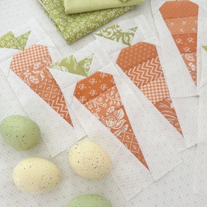 Puede incluir: Un conjunto de cinco formas de zanahoria de tela con fondo blanco y tela estampada en marrón y blanco. Las zanahorias de tela están dispuestas en forma de abanico con las puntas de las zanahorias hacia afuera. Hay tres huevos moteados en el fondo.