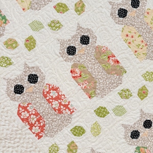 Peut inclure: Un tissu matelassé présentant un motif répétitif de formes de hiboux. Les hiboux sont fabriqués à partir de tissu beige avec des imprimés floraux rouges, roses et verts, et ont des yeux circulaires noirs. De petits hexagones verts et beiges sont dispersés.