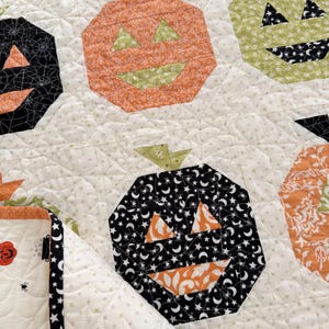 Puede incluir: Una tela acolchada con un patrón repetitivo de calabazas de Halloween en naranja, negro y verde claro. Las calabazas tienen ojos triangulares y bocas anchas, sobre un fondo crema con un diseño de costuras de estrellas y telarañas.