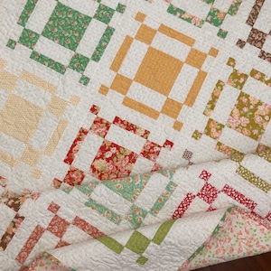 May include: A patchwork quilt featuring a repeating block pattern with a white background fabric printed with small dots. The blocks contain floral and solid fabric squares in shades of green, mustard yellow, red, and brown, with visible stitching.