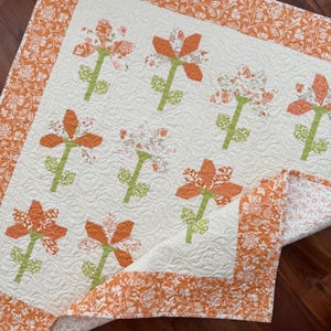 Puede incluir: Una manta acolchada con un centro color crema y un patrón repetitivo de flores estampadas en naranja y blanco sobre tallos verdes. La colcha está bordeada por un marco ancho de tela floral naranja y blanca con costuras detalladas.