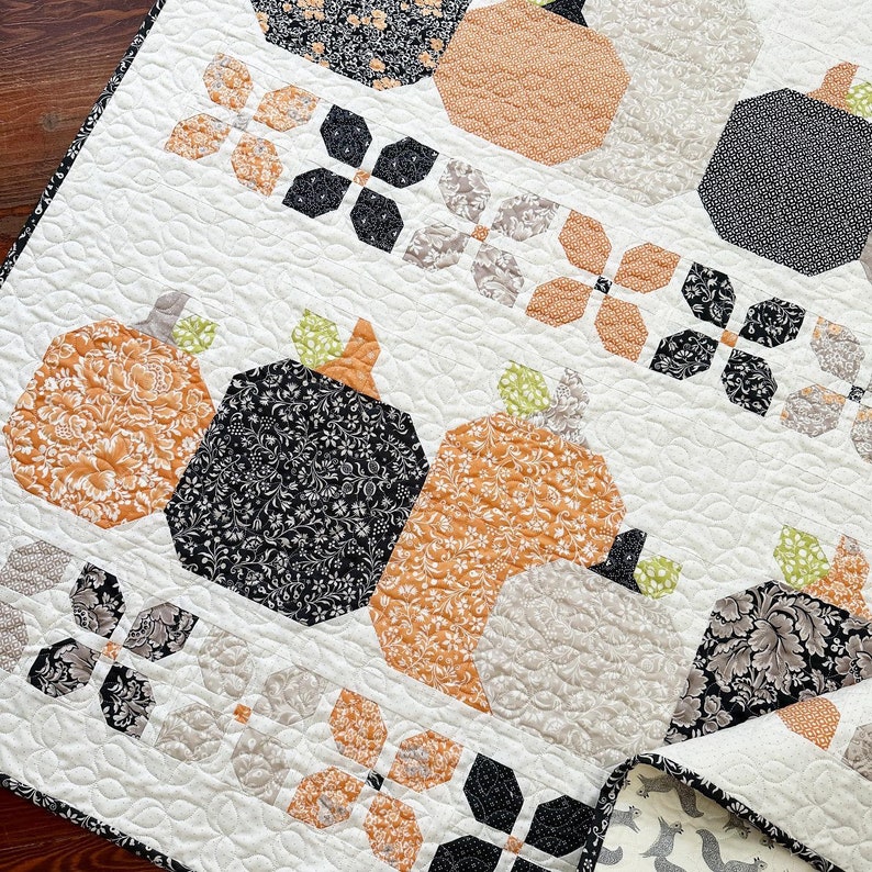 May include: A white quilt with a patchwork design featuring pumpkins, flowers, and leaves in shades of orange, black, and gray.