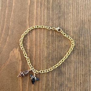 May include: Gold-toned chain bracelet with a seashell and a dark blue shark tooth charm. The bracelet is displayed on a wooden surface. The bracelet has a lobster claw clasp.