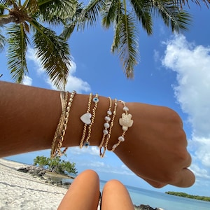 May include: A collection of gold bracelets adorns an arm against a backdrop of a sunny beach and palm trees. The bracelets include beaded, chain, and charm styles, featuring heart, pearl, and turtle designs. The sky is a bright blue with fluffy white clouds.