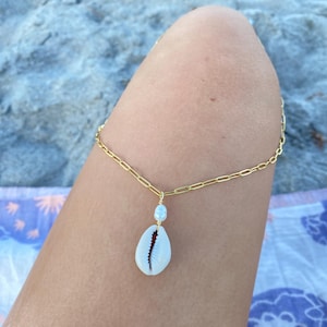 May include: A gold-colored chain anklet with a white seashell pendant and a small blue bead. The anklet is worn on a leg, with a beach scene in the background. The seashell has a dark interior.