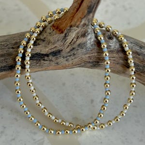 May include: Two beaded bracelets with gold, light blue, and white beads. The bracelets are draped over a piece of driftwood. The gold beads are larger than the light blue and white beads.