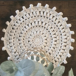 May include: A cream-coloured, crocheted doily with a circular design and scalloped edge. A decorative glass sphere with a woven pattern is partially visible beneath the doily. Eucalyptus leaves and sprigs add a natural touch.