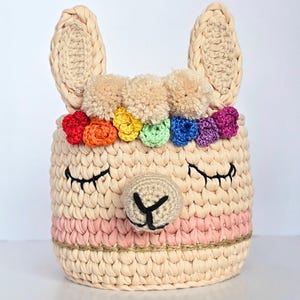 May include: A beige crochet llama basket with a pink stripe and a rainbow flower crown. The basket has closed eyes and a small nose. The ears are beige and stand up. The basket is round and has a woven texture.