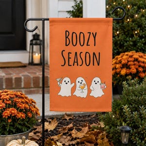 May include: A rectangular orange garden flag featuring the black text BOOZY SEASON above three white ghost illustrations, each holding a different cocktail glass. The flag is suspended from a black metal stand.