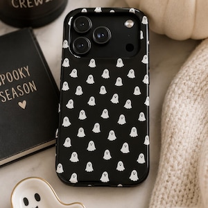 Op de afbeelding: Een zwarte smartphonehoes met een herhalend patroon van kleine witte spookjes. De hoes heeft een rechthoekige vorm met afgeronde hoeken en een uitsparing aan de bovenzijde voor de cameralenzen en de flitser van het toestel.