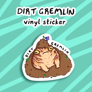 May include: A vinyl sticker featuring a cartoon illustration of a brown frog with yellow eyes, partially buried in dirt with a purple flower growing out of the dirt. The sticker has the words "Dirt Gremlin" written above the frog and "Dirt" and "Gremlin" written on either side of the frog.