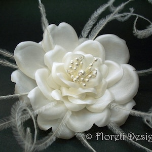 May include: Ivory fabric flower with delicate petals and a cluster of silver beads at the center. Wispy white feathers extend from the flower's edges. The design is set against a dark background.