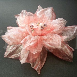 May include: A delicate, light pink fabric flower with multiple petals and a shimmering, metallic gold underside. The center of the flower is adorned with small, sparkling crystals. This floral accessory is suitable for weddings or special occasions.