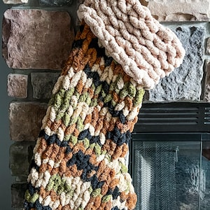 May include: A chunky knit Christmas stocking featuring a camouflage pattern in shades of olive green, brown, black, and cream. The stocking has a thick, folded cuff made of solid cream-colored yarn, creating a textured, soft appearance throughout.