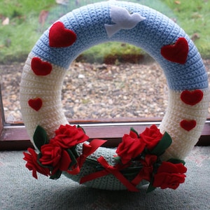 May include: A crocheted wreath with a blue and white pattern and red hearts. The wreath is decorated with red roses.