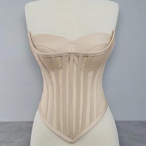 Op de afbeelding: Een beige korset op een mannequin. Het strapless ontwerp heeft verticale baleinen en een sweetheart halslijn. Het korset is gemaakt van een lichte, neutrale stof, waardoor een gestroomlijnd silhouet ontstaat. Het kledingstuk is ontworpen om de taille te accentueren en te ondersteunen.