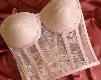 May include: A blush-colored corset with a sweetheart neckline. The bodice is made of a delicate lace fabric, with vertical boning and a sheer mesh panel. The corset is a lingerie item.