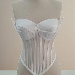 Puede incluir: Un cors&eacute; blanco exhibido en un maniqu&iacute;. El cors&eacute; presenta un corpi&ntilde;o estructurado con ballenas verticales y un escote coraz&oacute;n. El dise&ntilde;o incluye un panel transparente en la secci&oacute;n media y un dobladillo puntiagudo.