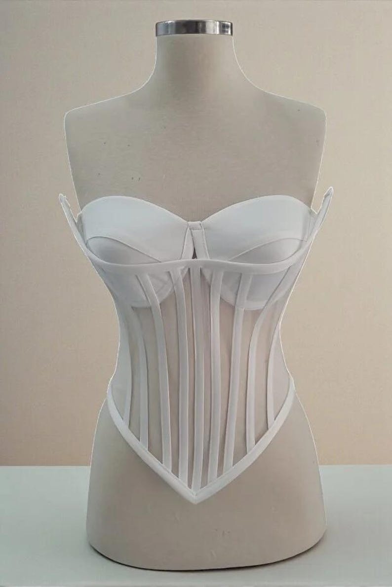 Puede incluir: Un cors&eacute; blanco exhibido en un maniqu&iacute;. El cors&eacute; presenta un corpi&ntilde;o estructurado con ballenas verticales y un escote coraz&oacute;n. El dise&ntilde;o incluye un panel transparente en la secci&oacute;n media y un dobladillo puntiagudo.