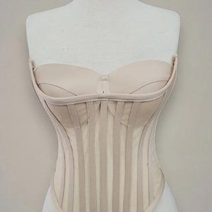 Puede incluir: Un cors&eacute; beige exhibido en un maniqu&iacute;. El corpi&ntilde;o sin tirantes presenta ballenas verticales y un escote coraz&oacute;n. El cors&eacute; est&aacute; hecho de una tela de color claro y tiene un dise&ntilde;o estructurado y ajustado. La est&eacute;tica general es de inspiraci&oacute;n vintage.