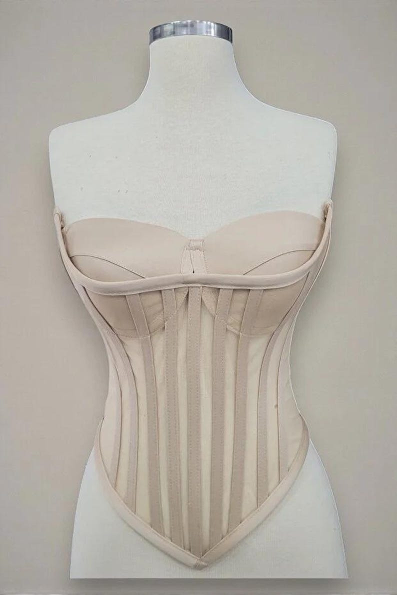 Puede incluir: Un cors&eacute; beige exhibido en un maniqu&iacute;. El corpi&ntilde;o sin tirantes presenta ballenas verticales y un escote coraz&oacute;n. El cors&eacute; est&aacute; hecho de una tela de color claro y tiene un dise&ntilde;o estructurado y ajustado. La est&eacute;tica general es de inspiraci&oacute;n vintage.
