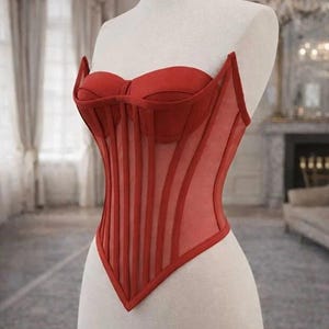 Puede incluir: Un cors&eacute; rojo exhibido en un maniqu&iacute;. El cors&eacute; presenta un corpi&ntilde;o estructurado con ballenas verticales y un escote coraz&oacute;n. El dise&ntilde;o incluye paneles transparentes y un dobladillo en punta, mostrando una mezcla de estructura y detalles delicados.