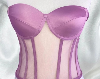 May include: A strapless lavender corset with a sweetheart neckline. The bodice features a satin finish on the cups and vertical sheer panels. The corset is designed to cinch the waist and create a flattering silhouette.