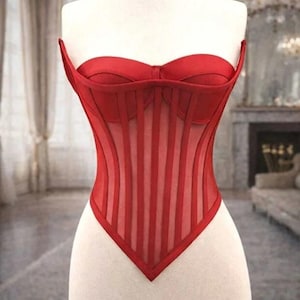 Puede incluir: Un cors&eacute; rojo exhibido en un maniqu&iacute;. El cors&eacute; presenta un corpi&ntilde;o estructurado con paneles verticales transparentes y un escote coraz&oacute;n. La prenda est&aacute; hecha de tela roja y est&aacute; dise&ntilde;ada para ce&ntilde;ir la cintura.