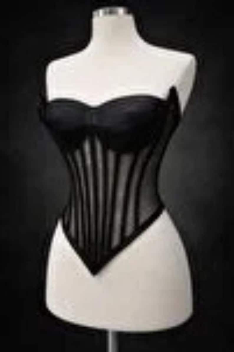 Puede incluir: Un cors&eacute; negro exhibido en un maniqu&iacute;. El cors&eacute; presenta un escote coraz&oacute;n y ballenas verticales, creando una silueta estructurada. La prenda est&aacute; hecha de una combinaci&oacute;n de materiales opacos y transparentes.