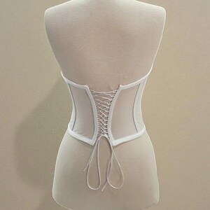 Puede incluir: Un cors&eacute; blanco con la espalda con cordones, exhibido en un maniqu&iacute;. El cors&eacute; tiene un dise&ntilde;o estructurado con un panel transparente en la espalda. La prenda est&aacute; hecha de una tela de color claro.