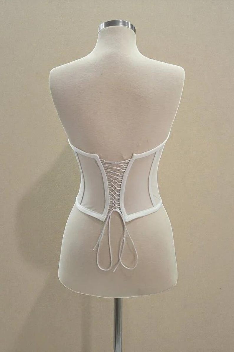 Puede incluir: Un cors&eacute; blanco con la espalda con cordones, exhibido en un maniqu&iacute;. El cors&eacute; tiene un dise&ntilde;o estructurado con un panel transparente en la espalda. La prenda est&aacute; hecha de una tela de color claro.