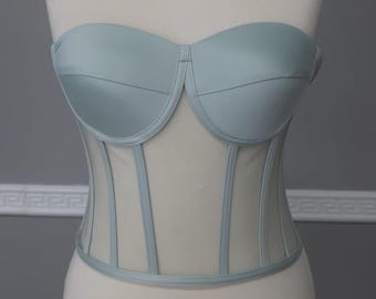 May include: A light blue strapless corset top with a sweetheart neckline. The bodice features a structured design with vertical boning and a sheer mesh panel. The corset is displayed on a mannequin, showcasing its form and details.
