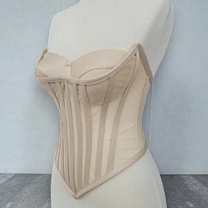 Puede incluir: Un cors&eacute; beige expuesto en un maniqu&iacute;. El corpi&ntilde;o sin tirantes presenta ballenas verticales y un escote coraz&oacute;n. El cors&eacute; est&aacute; hecho de una tela transparente, similar a una malla, con paneles y ribetes opacos.