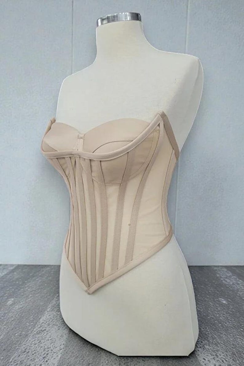 Puede incluir: Un cors&eacute; beige expuesto en un maniqu&iacute;. El corpi&ntilde;o sin tirantes presenta ballenas verticales y un escote coraz&oacute;n. El cors&eacute; est&aacute; hecho de una tela transparente, similar a una malla, con paneles y ribetes opacos.