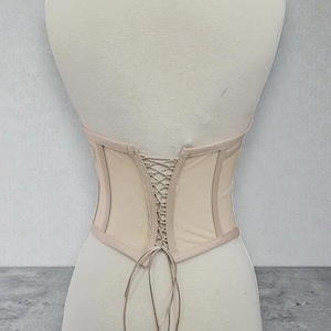 Puede incluir: Un cors&eacute; beige con la espalda con cordones se muestra en un maniqu&iacute;. El cors&eacute; tiene un dise&ntilde;o estructurado con una tela beige m&aacute;s clara y un ribete beige m&aacute;s oscuro. La parte trasera presenta un detalle de cordones entrecruzados y la parte inferior tiene forma curva.