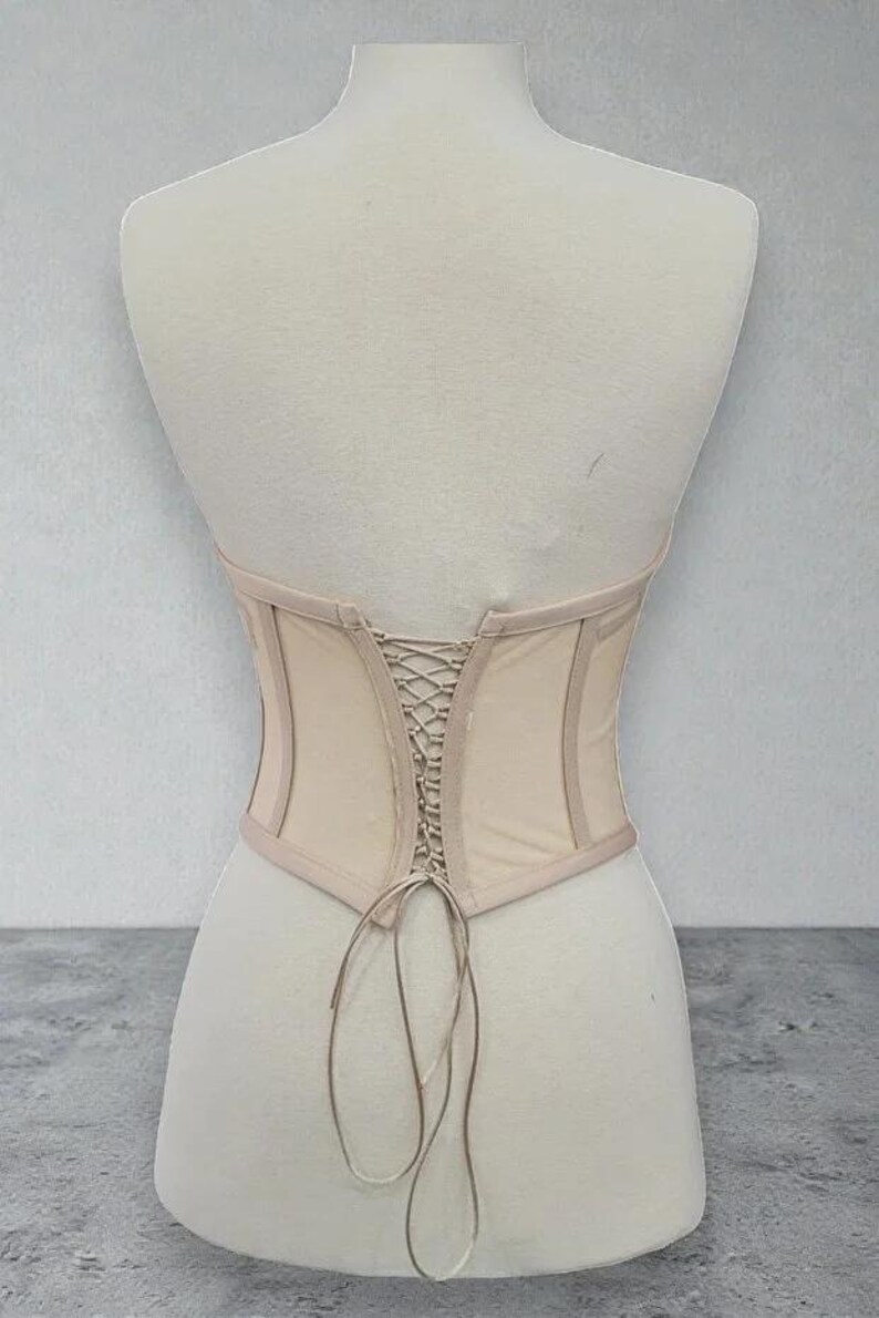 Puede incluir: Un cors&eacute; beige con la espalda con cordones se muestra en un maniqu&iacute;. El cors&eacute; tiene un dise&ntilde;o estructurado con una tela beige m&aacute;s clara y un ribete beige m&aacute;s oscuro. La parte trasera presenta un detalle de cordones entrecruzados y la parte inferior tiene forma curva.