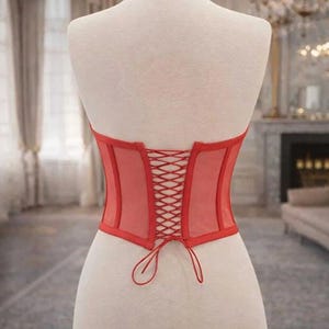 Puede incluir: Un cors&eacute; rojo exhibido en un maniqu&iacute;. El cors&eacute; presenta un dise&ntilde;o de malla transparente con una espalda con cordones. Los bordes est&aacute;n ribeteados con una tela roja lisa. El maniqu&iacute; se encuentra sobre un fondo borroso.