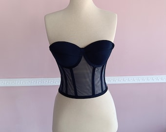 May include: A navy blue strapless corset displayed on a mannequin. The corset features a sheer mesh panel in the center, framed by solid navy fabric. The bust is structured with molded cups, and the garment has a smooth, tailored appearance.