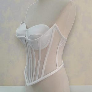 Puede incluir: Un cors&eacute; blanco exhibido en un maniqu&iacute;. El cors&eacute; presenta un corpi&ntilde;o estructurado con ballenas verticales y una superposici&oacute;n de malla transparente. El dise&ntilde;o incluye un escote coraz&oacute;n y una cintura definida.
