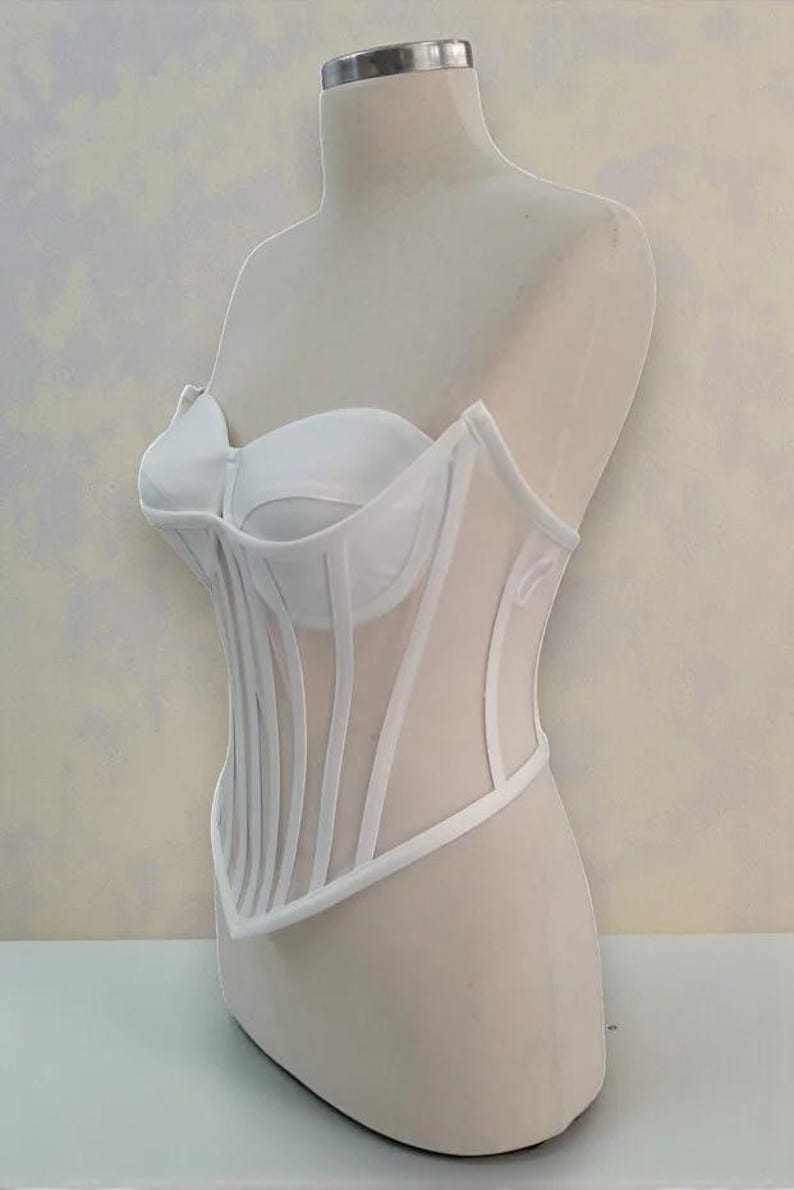Puede incluir: Un cors&eacute; blanco exhibido en un maniqu&iacute;. El cors&eacute; presenta un corpi&ntilde;o estructurado con ballenas verticales y una superposici&oacute;n de malla transparente. El dise&ntilde;o incluye un escote coraz&oacute;n y una cintura definida.