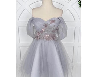 May include: A pale grey off-the-shoulder dress with a sweetheart neckline. The bodice features floral embroidery and beadwork. The short skirt is layered with tulle, and the sleeves are sheer. The dress is displayed on a mannequin.