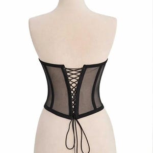 Puede incluir: Cors&eacute; negro con frente y espalda de malla transparente, exhibido en un maniqu&iacute;. El cors&eacute; presenta un dise&ntilde;o estructurado con espalda con cordones y ribete negro. La prenda est&aacute; dise&ntilde;ada para ce&ntilde;ir la cintura y crear una silueta definida.