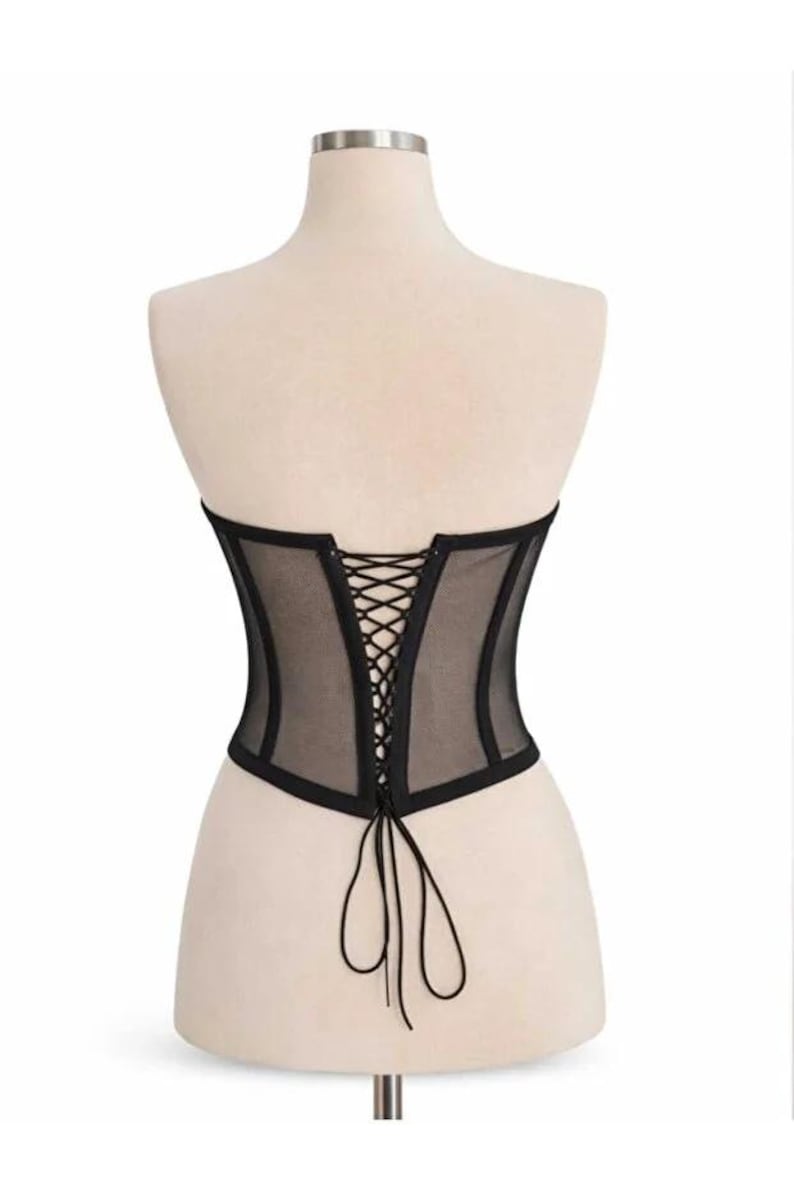 Puede incluir: Cors&eacute; negro con frente y espalda de malla transparente, exhibido en un maniqu&iacute;. El cors&eacute; presenta un dise&ntilde;o estructurado con espalda con cordones y ribete negro. La prenda est&aacute; dise&ntilde;ada para ce&ntilde;ir la cintura y crear una silueta definida.