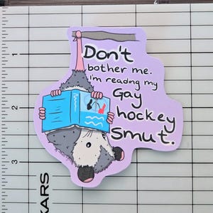 Peut inclure: Autocollant lavande avec un opossum de dessin animé lisant un livre. Le texte sur l'autocollant se lit comme suit : "Ne me dérangez pas. Je lis mon Gay hockey smut."