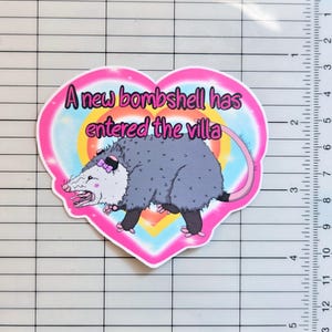 May include: A heart-shaped sticker featuring a grey opossum with a pink bow, set against a rainbow background with the text 'A new bombshell has entered the villa'. A ruler on the right shows the sticker is approximately 10 cm wide.