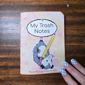Peut inclure: Un carnet intitulé "My Trash Notes" présente un opossum de dessin animé tenant un crayon. L'opossum est gris et blanc, avec des touches de rose. Le carnet a un fond coloré et le texte "Peaceful opossum art" en bas.